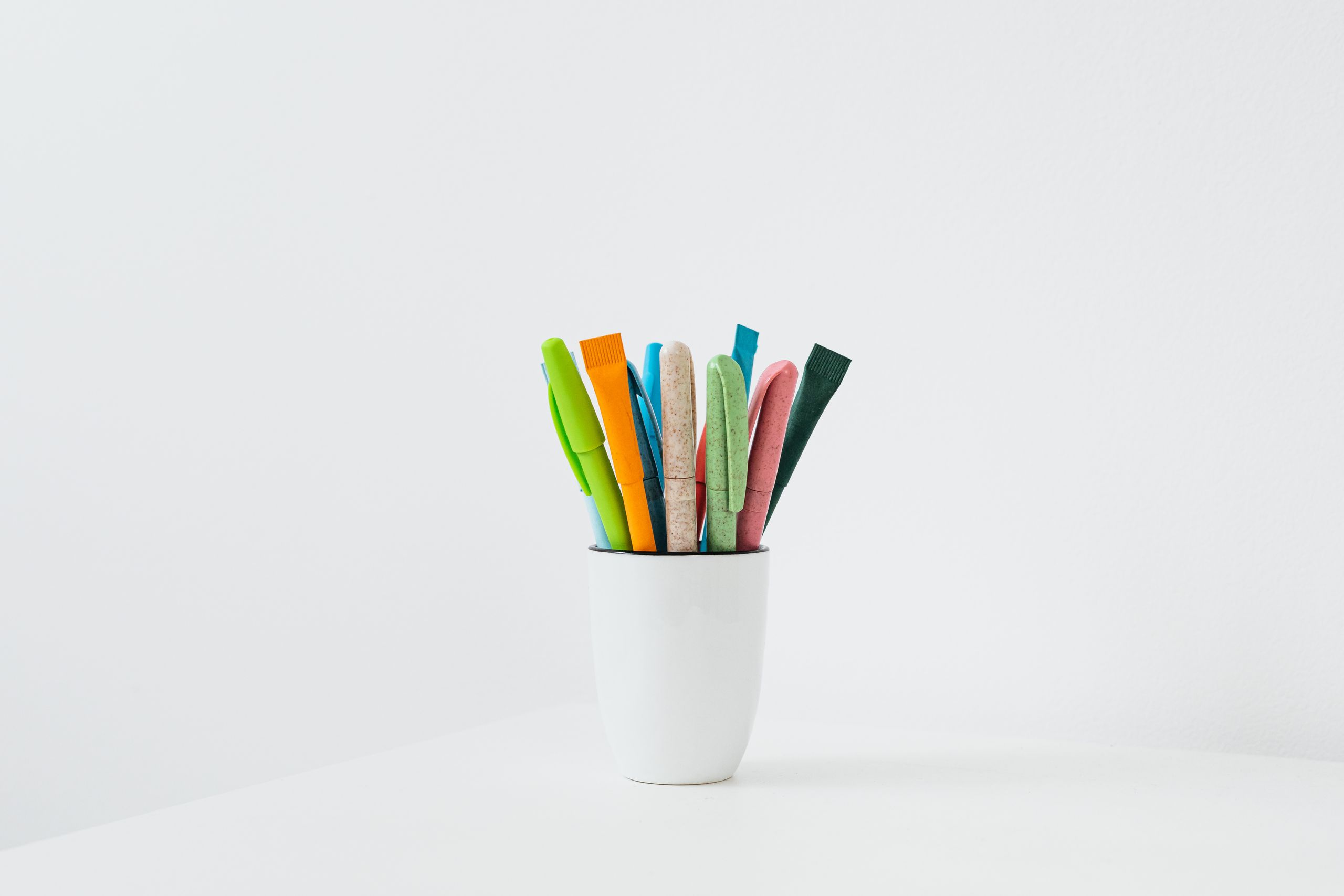Symbolbild. Weißer Becher auf einem weißen Tisch. In dem Becher stehen ca. elf bunte Stifte – jeweils mit einer Kappe darauf.